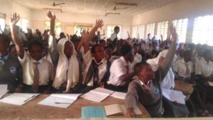 School girls seated and some raising hands, an indication of answering or asking a question.